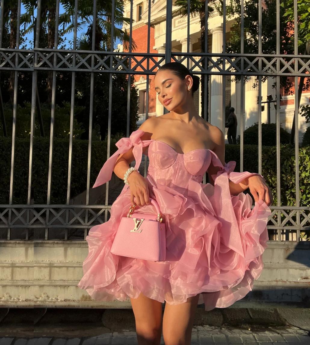 Pink organza mini dress with sweetheart neckline, tie straps, and voluminous ruffled skirt, styled with a matching handbag.