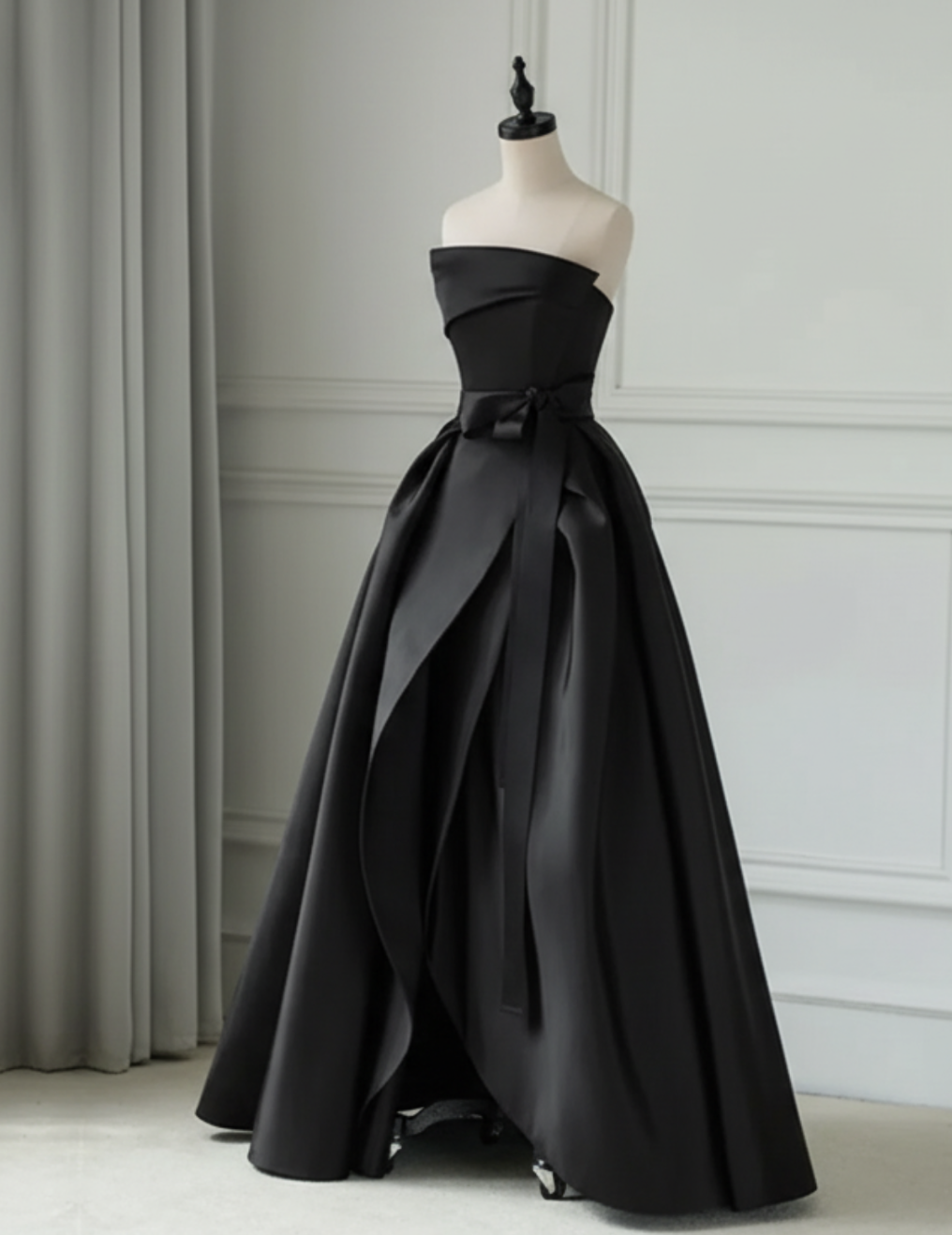 Black strapless satin twill gown with a pleated A-line silhouette, ruched bodice, and elegant waist sash on a mannequin.