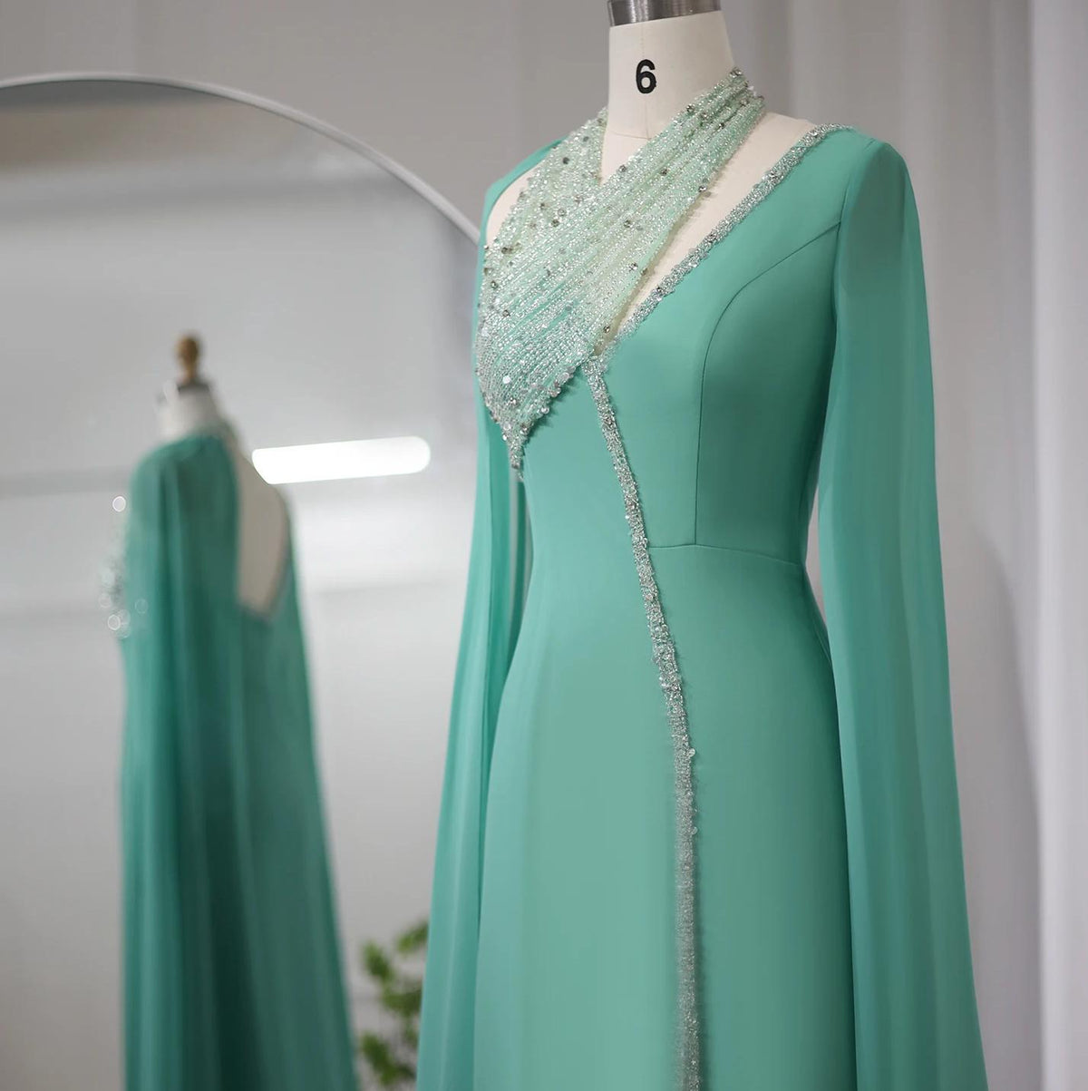 Teal chiffon Selma gown on a mannequin featuring intricate crystal beading, a halter neckline, and dramatic cape sleeves.
