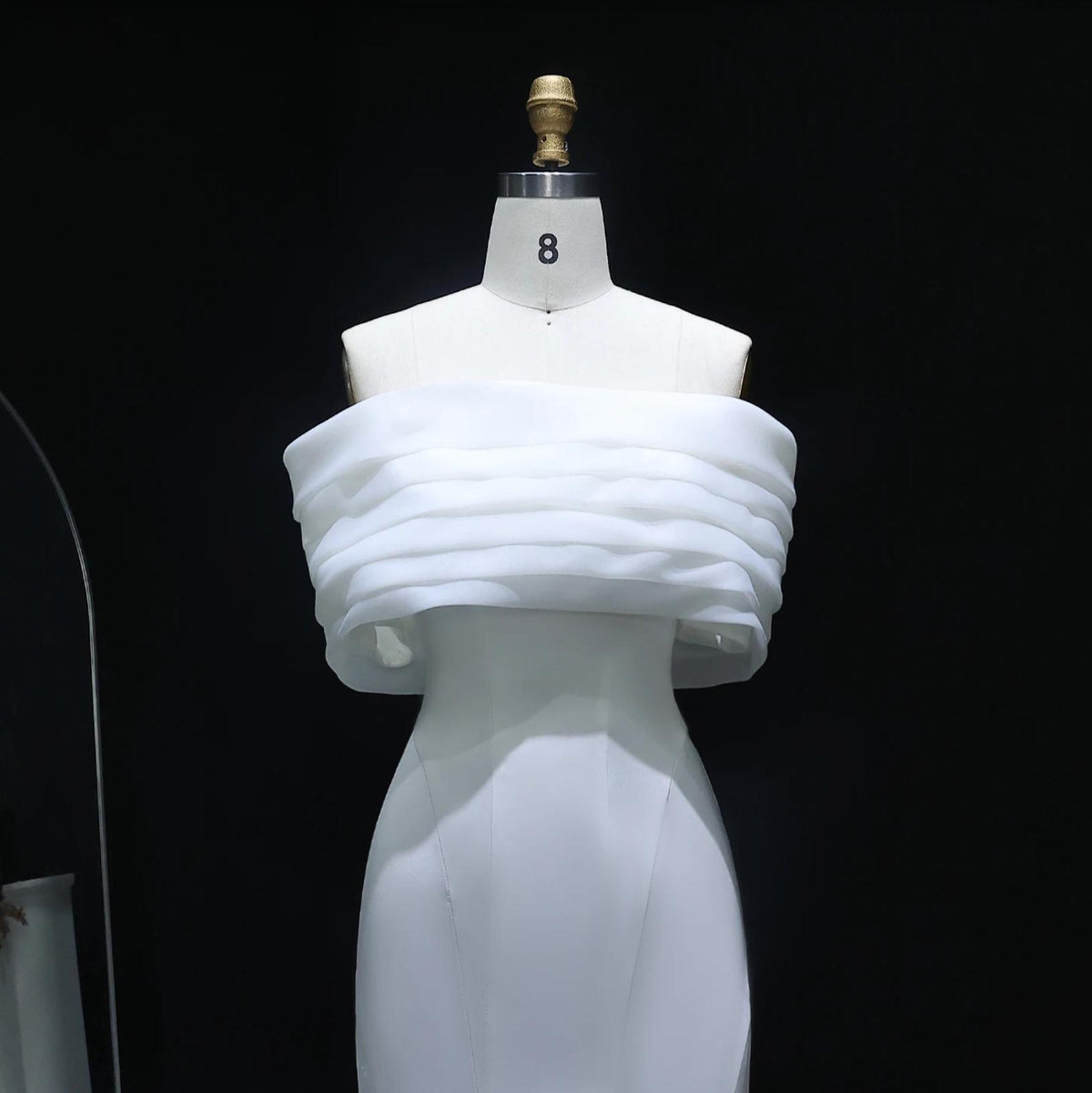 White off-shoulder mermaid evening gown with layered satin bodice on a mannequin against a black background.