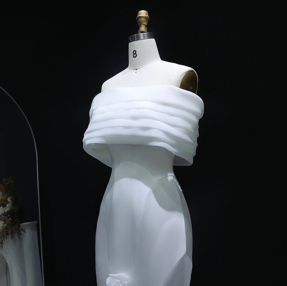 White off-shoulder mermaid gown on a mannequin, featuring layered satin fabric and delicate 3D floral accents.