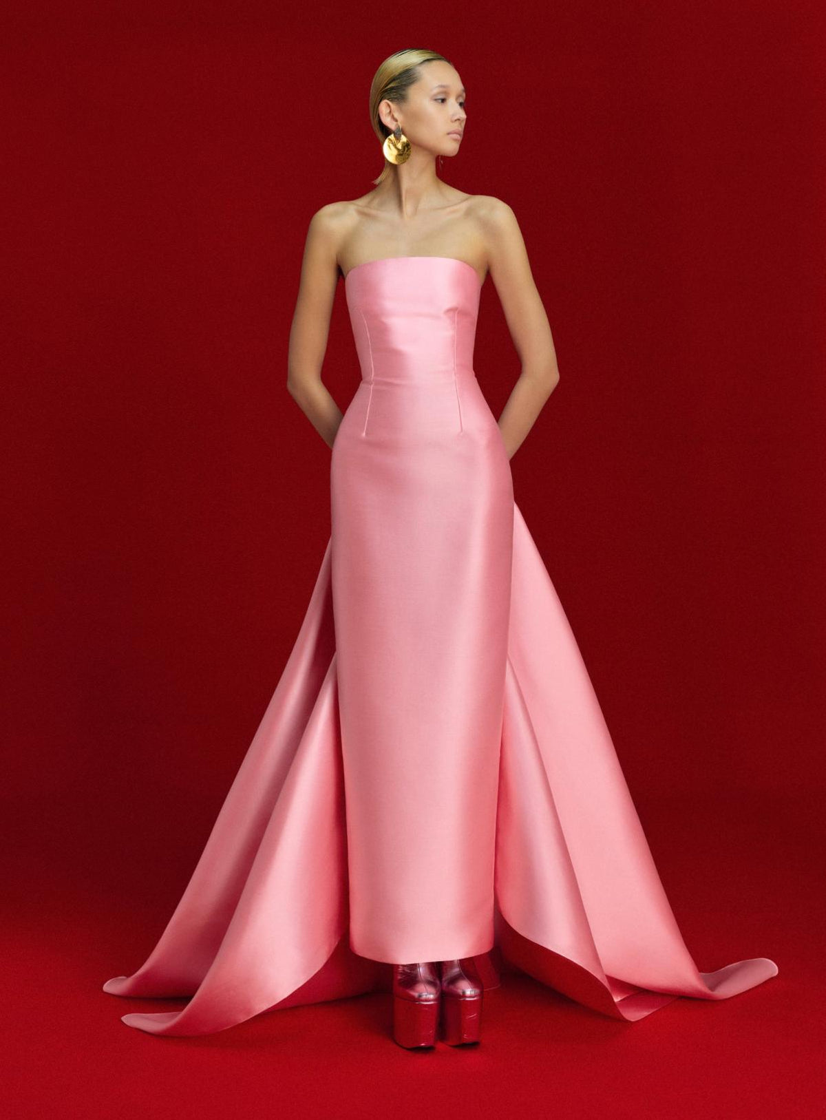 Strapless pink satin floor-length gown with a dramatic detachable train against a bold red background.