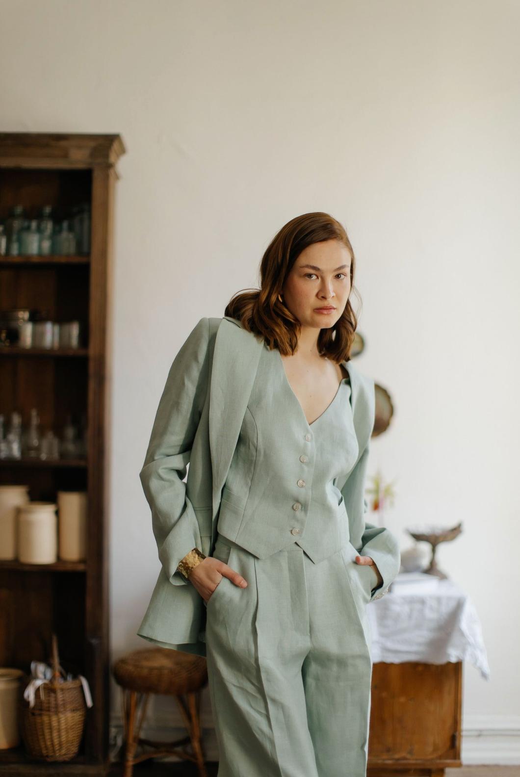 Woman wearing a sage green three-piece linen suit with a tailored jacket, button-up vest, and flowing palazzo pants.
