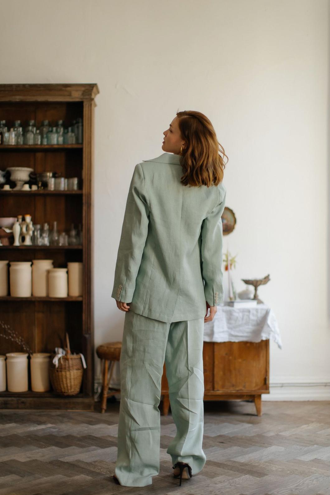 Woman wearing a sage green three-piece linen suit with a tailored jacket and flowing palazzo pants in a rustic room.