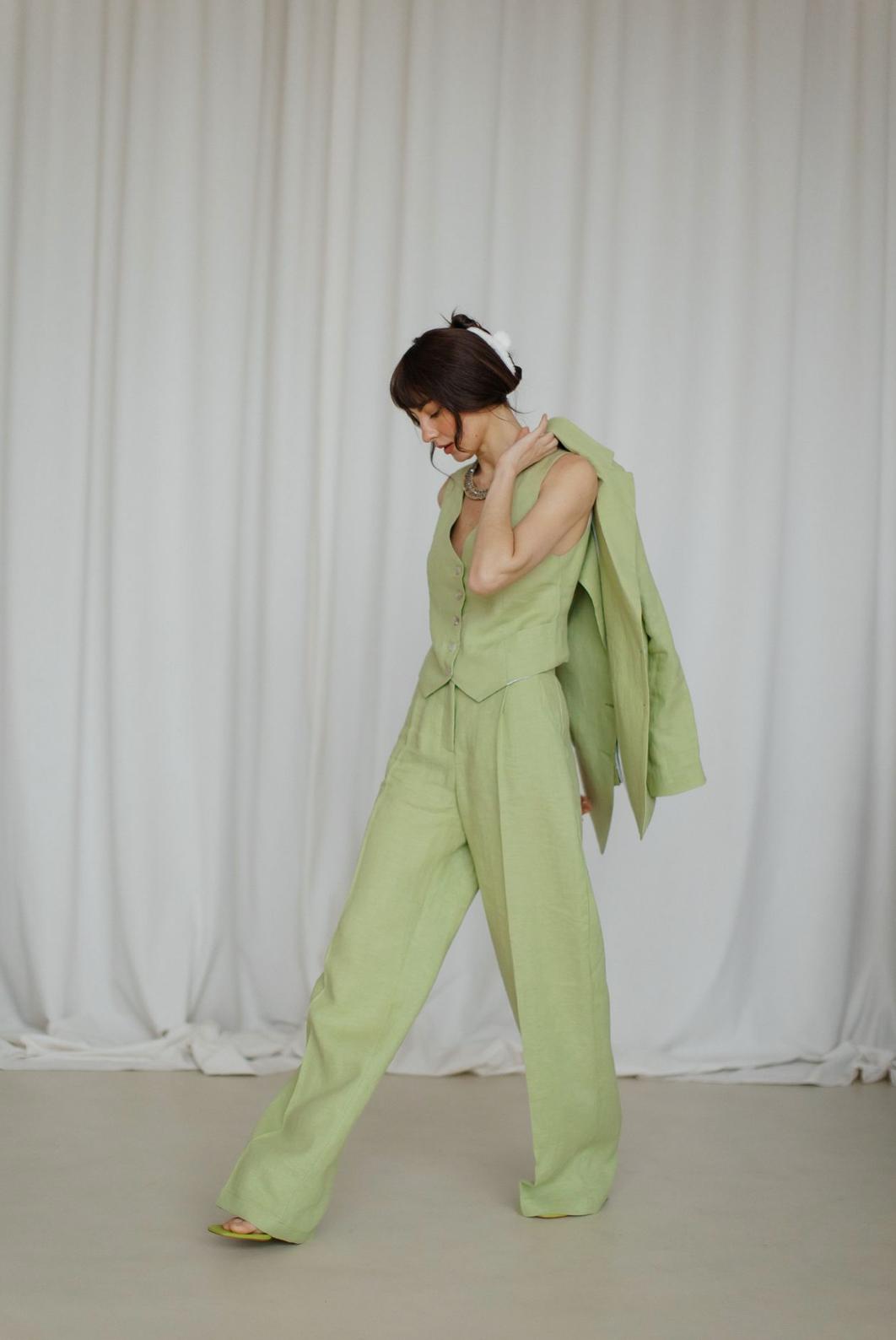 Woman wearing a sage green three-piece linen suit with a tailored vest, palazzo pants, and a jacket draped over her shoulder.