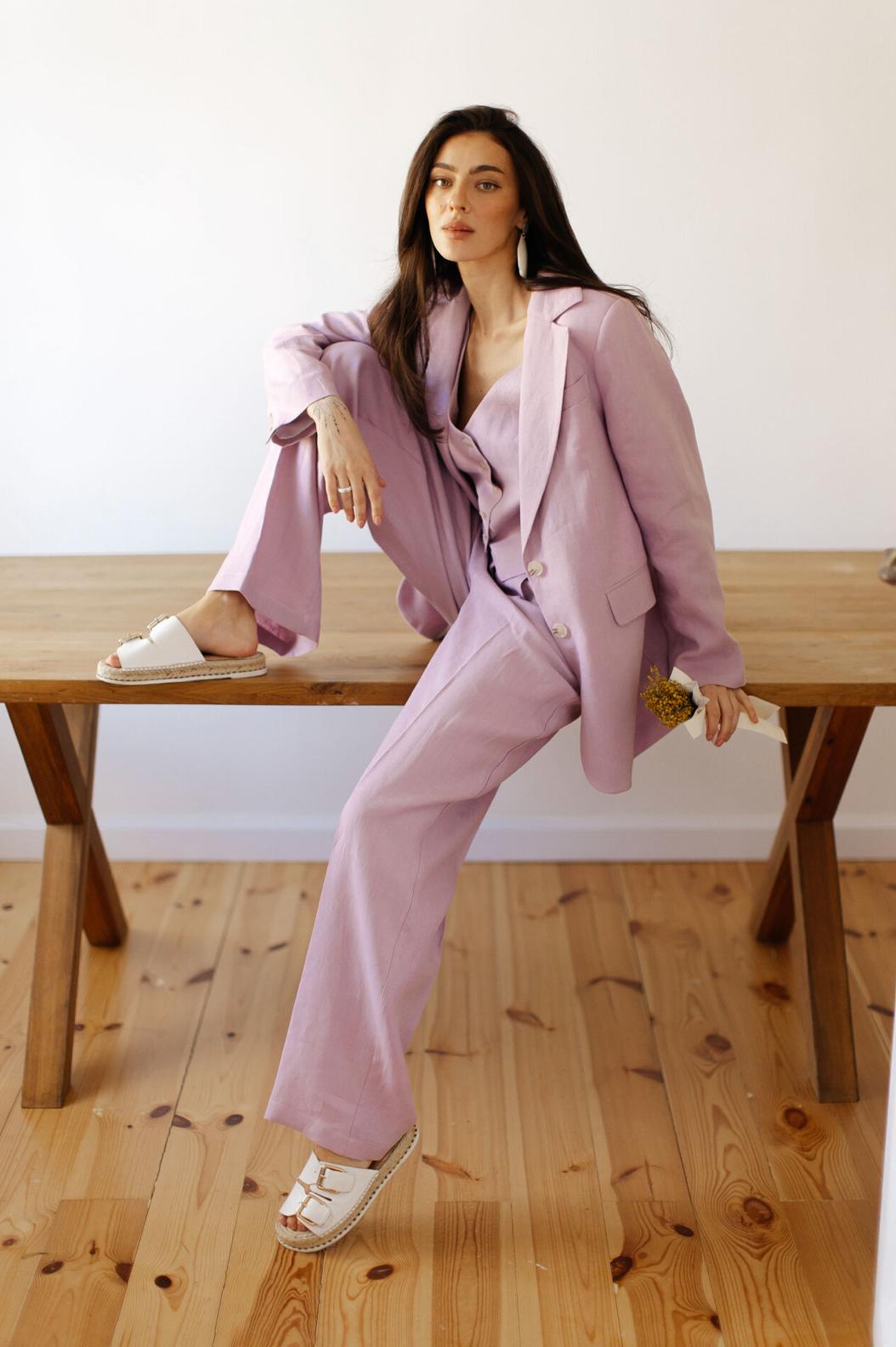 Woman wearing a lavender three-piece linen suit with a tailored jacket, vest, and palazzo pants, sitting on a wooden table.