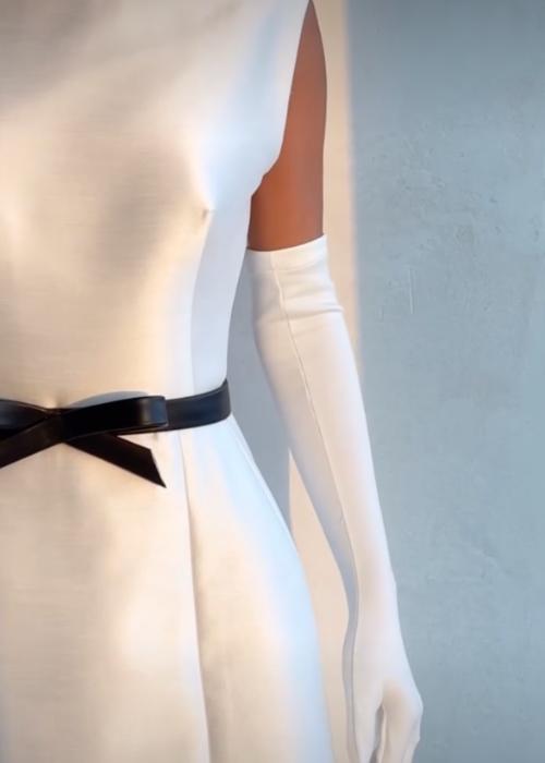 White satin mini dress with a sleeveless o-neckline, A-line silhouette, and a black bow belt, styled with long white gloves.