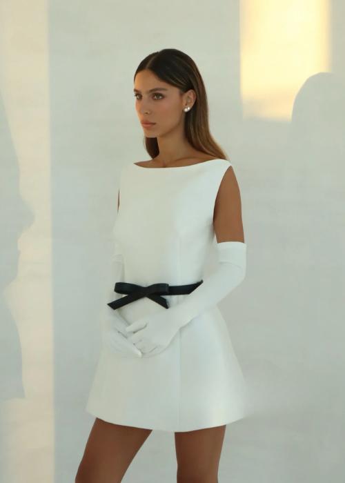 White satin mini dress with an o-neckline, a-line silhouette, and a black bow waist belt worn with matching opera gloves.