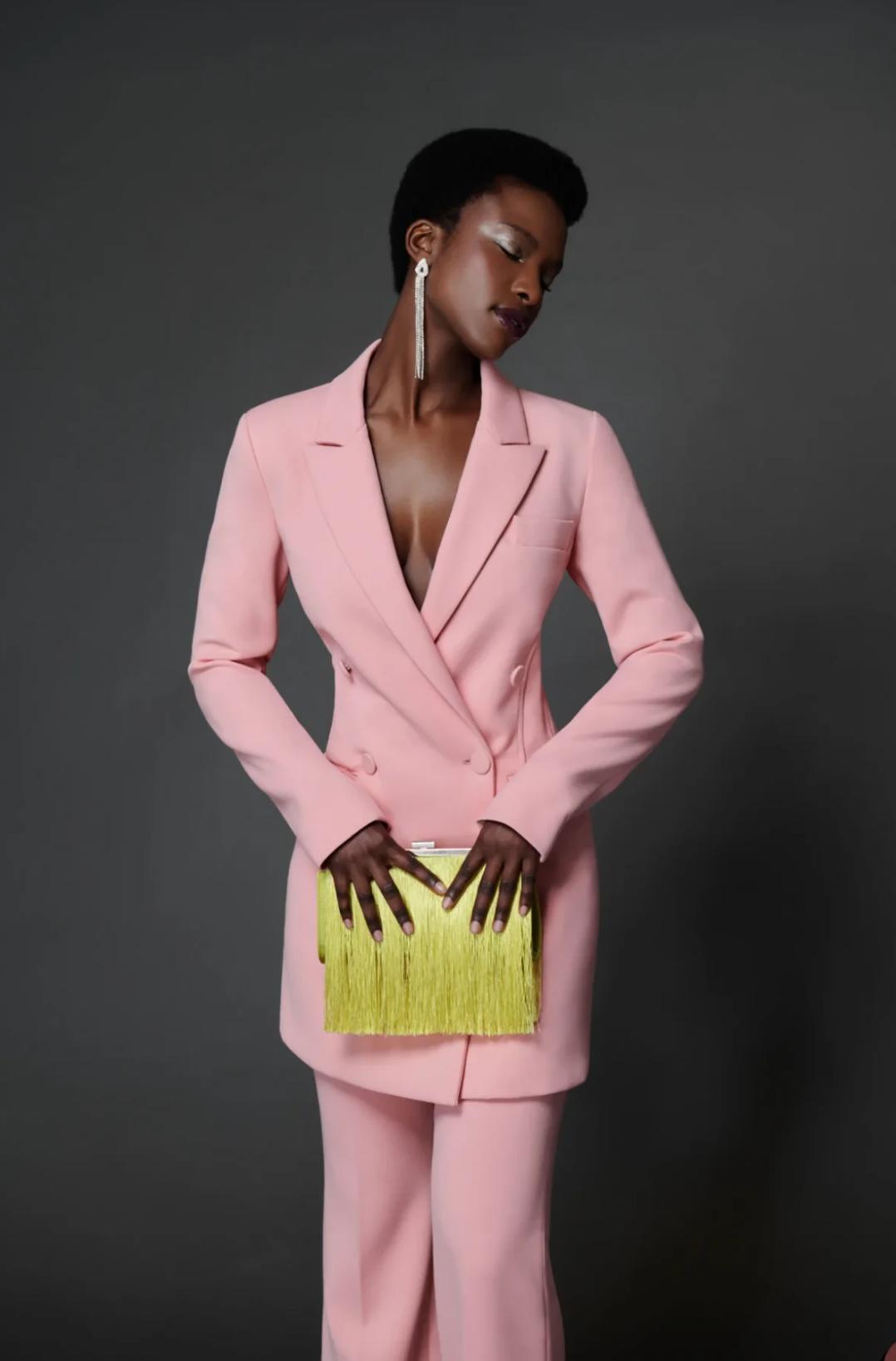 Model wearing a tailored pink satin double-breasted two-piece suit with a bright yellow fringed clutch.