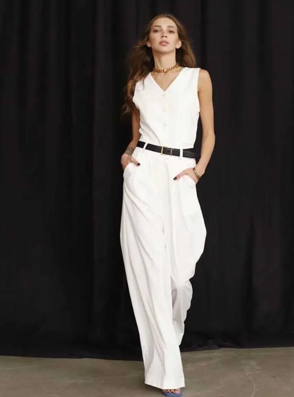 Woman wearing a white linen three-piece suit set with a buttoned vest and wide-leg trousers against a black background.