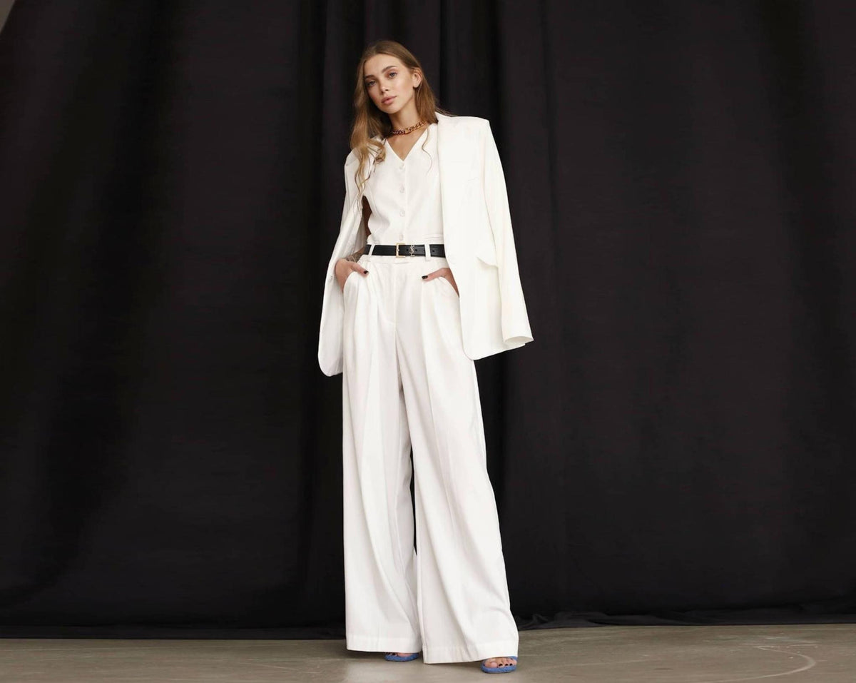 Woman wearing a white three-piece linen suit with a loose blazer, buttoned vest, and wide-leg trousers against black.