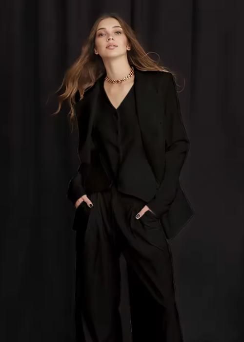 Woman wearing a black three-piece linen suit with a loose blazer, vest, and wide-leg trousers against a dark background.