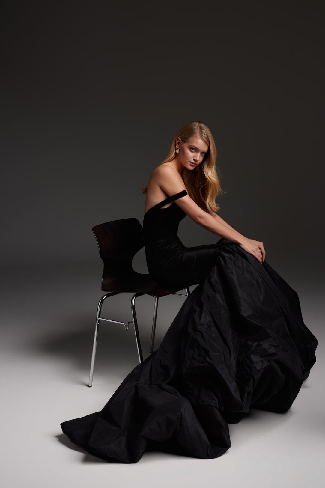Model wearing a black velvet and satin mermaid evening gown with a square neckline, seated on a chair.