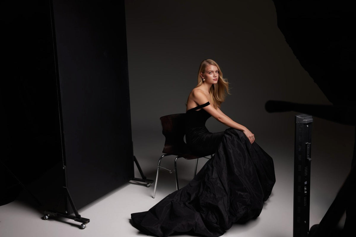 Model wearing a black satin mermaid gown with a square neckline and flowing skirt in a studio setting.
