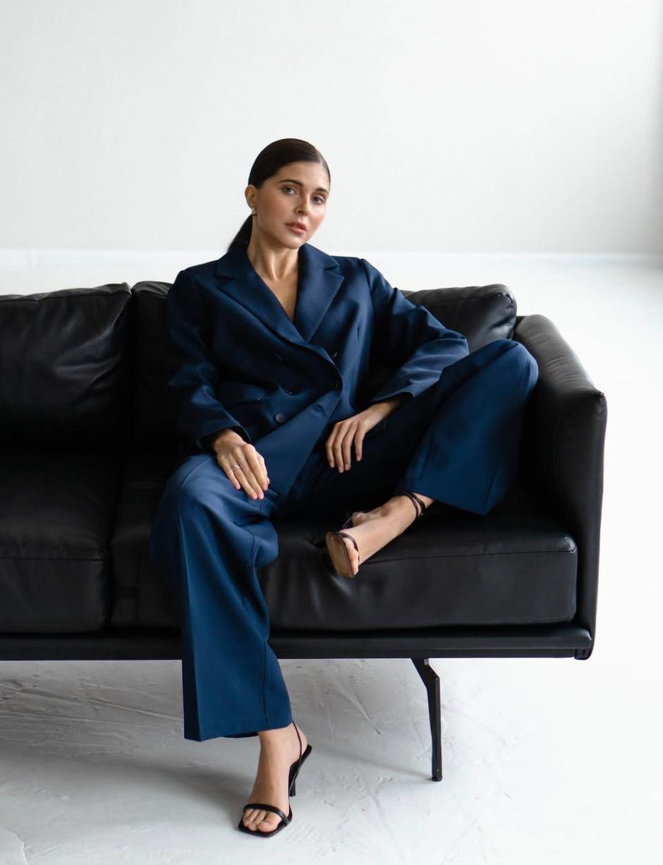 Woman wearing a dark blue double-breasted serge suit with ankle-length trousers, sitting on a black leather sofa.