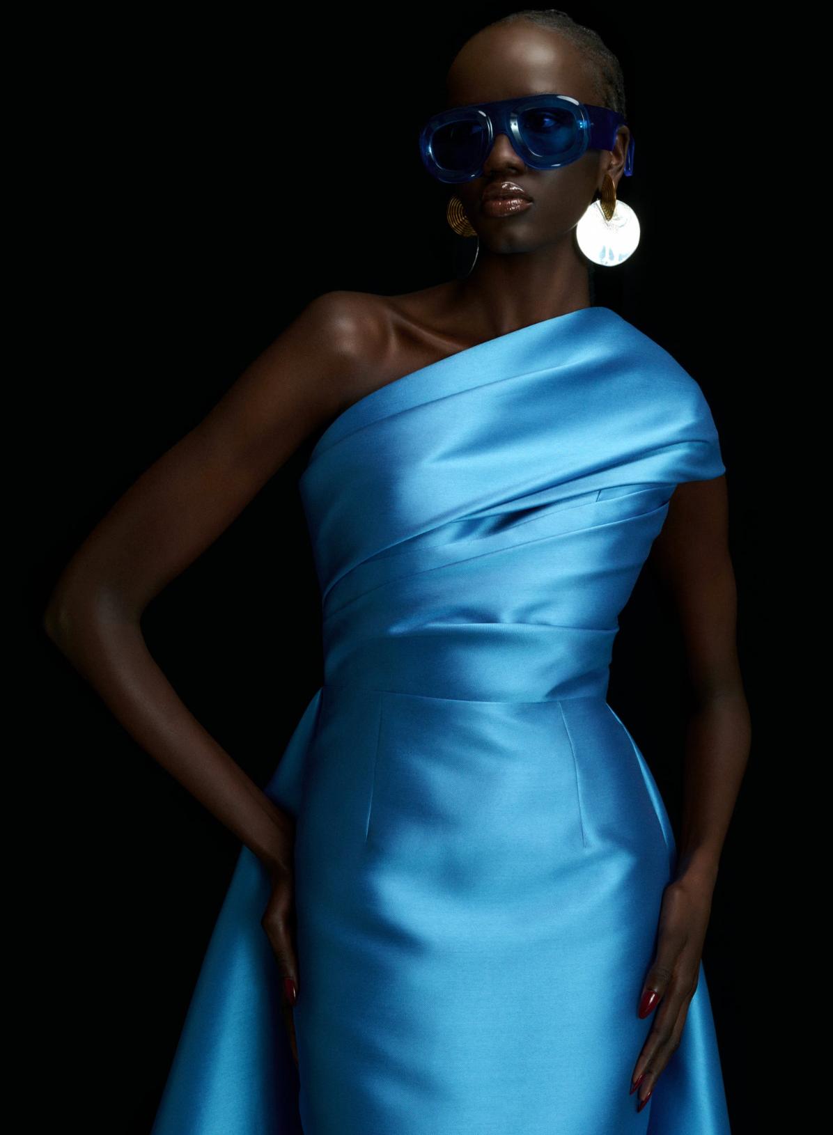 Model wearing a bright blue satin one-shoulder gown with pleated bodice and A-line skirt against a black background.