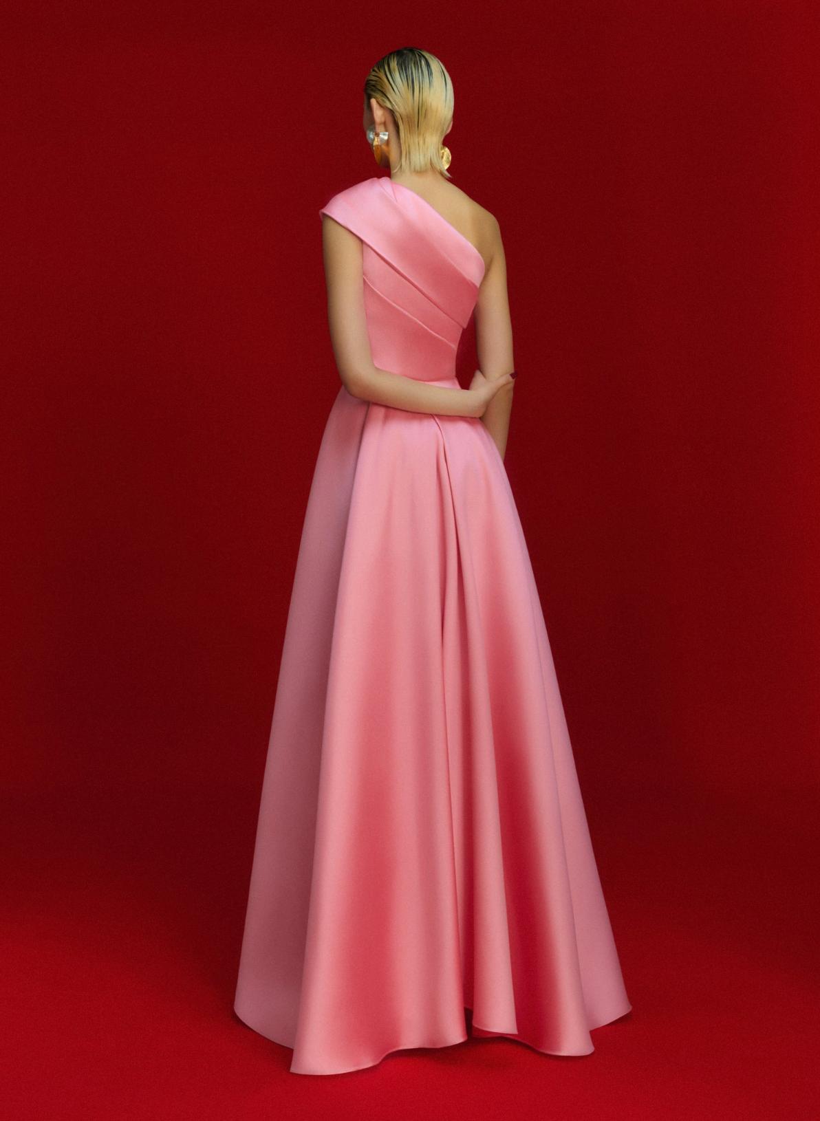 Pink satin one-shoulder A-line gown with a fitted waist and pleated skirt against a red background.